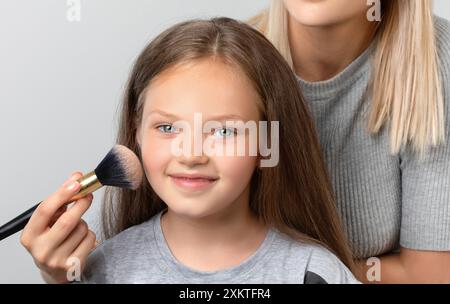 Portrait d'une belle fille souriante avec les yeux bleus avec les cheveux brun clair, maquilleuse applique de la poudre pour bébé sur son visage. Maquillage et cosmet pour enfants Banque D'Images