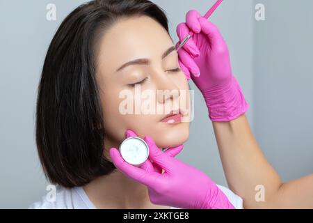 Fille adolescente Brunette ayant un tatouage de maquillage permanent sur ses sourcils. Le maquilleur fait des marques avec de la pâte blanche pour le tatouage des sourcils. Professio Banque D'Images