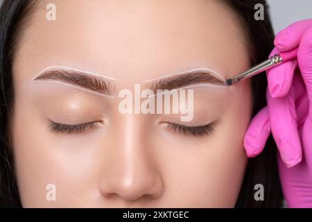 Fille adolescente Brunette ayant un tatouage de maquillage permanent sur ses sourcils. Le maquilleur fait des marques avec de la pâte blanche pour le tatouage des sourcils. Professio Banque D'Images