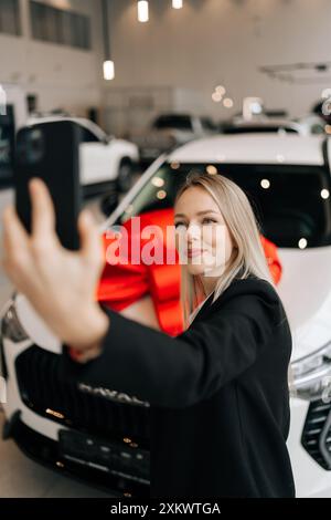 Plan vertical de la cliente féminine heureuse en costume noir faisant selfie shot pov sur téléphone mobile choisir auto veulent acheter une nouvelle automobile dans la salle d'exposition de voiture. Banque D'Images