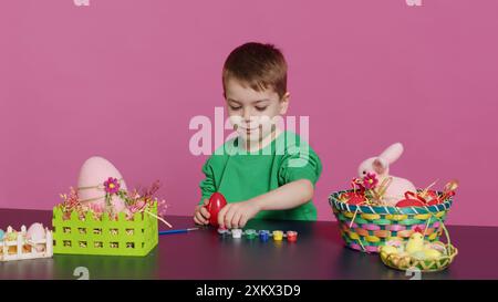 Petit enfant doux décorant des oeufs avec des timbres et de l'aquarelle, préparation pour les vacances du dimanche de pâques. Joyeux petit garçon ayant du plaisir de colorer des décorations festives, peindre des dessins en studio. Caméra A. Banque D'Images