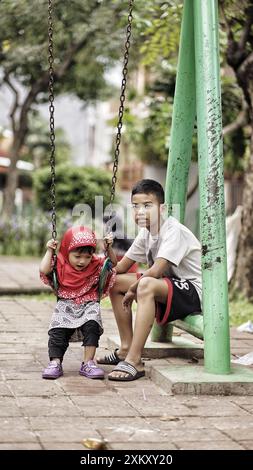 un garçon s'occupe de sa petite sœur qui joue sur une balançoire dans un parc pendant la journée Banque D'Images