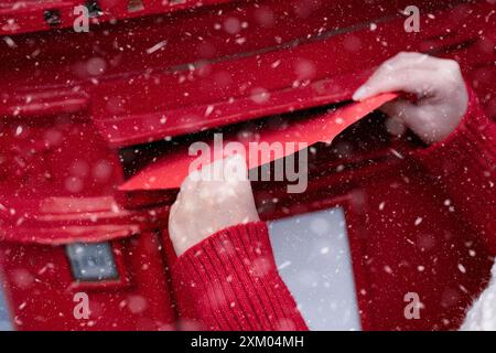 Femme dans un manteau rouge et chapeau blanc mettant une carte dans la boîte aux lettres rouge et se promenant dans une ville anglaise un jour neigeux avant Noël Banque D'Images