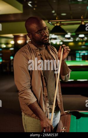 Jeune homme afro-américain à la craie de snooker frottant la pointe de queue de billard pour l'empêcher de glisser avant de jouer au billard dans la salle de jeux Banque D'Images