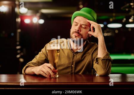 Jeune homme pensif en chapeau vert regardant un verre de bière tout en étant assis près du comptoir du bar dans un pub irlandais et célébrant la Saint Patrick Banque D'Images