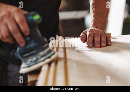 Menuisier qualifié ponçant le bois avec une ponceuse électrique en atelier, créant de la sciure de bois. Artisanat et dévouement montré dans la fabrication de meubles faits à la main Banque D'Images