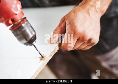 Le menuisier concentré perce un trou dans une planche blanche, créant de la sciure de bois. Dévouement et précision sont essentiels à son métier Banque D'Images