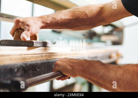 Gros plan des mains d'un menuisier à l'aide d'un rabot à main pour lisser une planche de bois dans un atelier de menuiserie, démontrant les techniques traditionnelles de travail du bois Banque D'Images