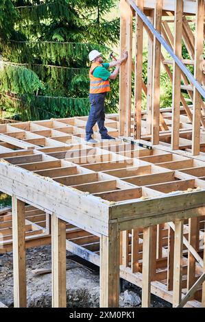 Charpentier construisant une maison à ossature en bois de deux étages près de la forêt. Homme barbu martelant des clous dans la structure, portant un casque de protection, gilet de construction. Concept de construction écologique moderne Banque D'Images