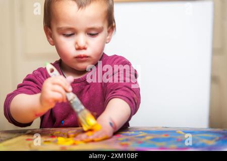 Les pinceaux pour enfant en bas âge peignent de la peinture dorée sur sa main. Derrière lui se trouve une toile vierge. Banque D'Images