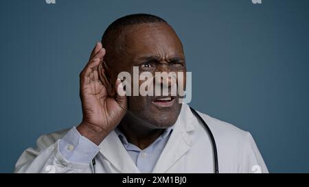 Gros plan confus homme afro-américain perplexe médecin masculin dans un fond bleu studio tenant la main près de l'oreille problème de perte auditive pencher la caméra vers l'avant Banque D'Images