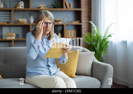 Femme senior assise sur le canapé regardant inquiet après avoir reçu une lettre importante. Femme lisant un document avec une expression préoccupée. Concept d'anxiété, de mauvaises nouvelles, de finances et de factures inattendues Banque D'Images