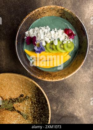 Bol à smoothie spiruline avec kiwi de mangue et flocons de noix de coco, joliment décoré. Banque D'Images