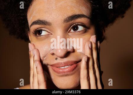 Une jeune femme à la peau foncée applique doucement un produit nettoyant sur son visage, mettant en valeur la beauté de la diversité. Banque D'Images