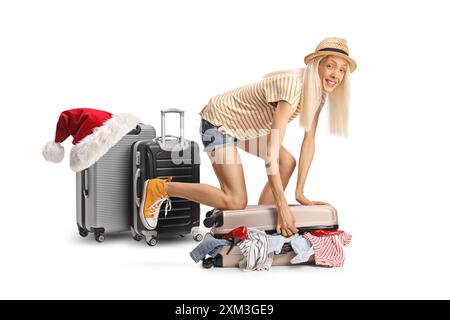 Jeune femme se préparant pour un voyage de vacances de noël et essayant de fermer la valise pleine de vêtements isolés sur fond blanc Banque D'Images