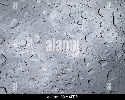 Vue rapprochée de gouttes de pluie sur verre, capturant des motifs complexes et des reflets sur un ciel nuageux et couvert, mettant en valeur la beauté des éléments naturels dans les environnements urbains. Banque D'Images