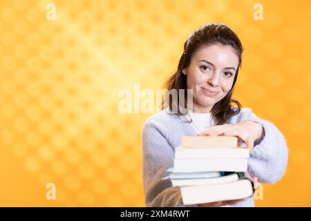 Femme tenant une pile de livres, pointant vers la caméra, isolée sur fond jaune de studio. Portrait de dame souriante de Bookworm présentant une pile de romans, exhortant les gens à lire Banque D'Images
