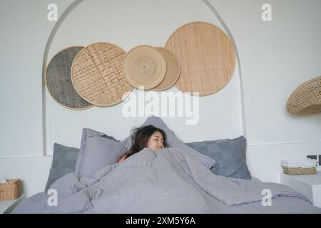 Femme asiatique gardant les yeux fermés et couvrant le corps avec une couverture grise, dormant sur le lit dans la chambre. Concept relaxant de vacances. Banque D'Images