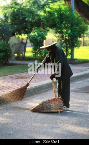 Un travailleur du parc portant un chapeau à large bord et un uniforme bleu balayant les feuilles mortes avec un balai en bambou dans un plateau de pelle sur un chemin pavé dans Lumpini Park, Bangkok, Thaïlande. Banque D'Images