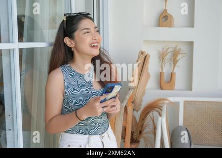 Portrait d'une jeune femme asiatique joyeuse debout à la terrasse, souriante et tenant un smartphone tout en portant une tenue décontractée en vacances Banque D'Images