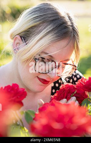 Une femme blonde dans une robe noire avec des pois blancs apprécie les roses d'été Banque D'Images