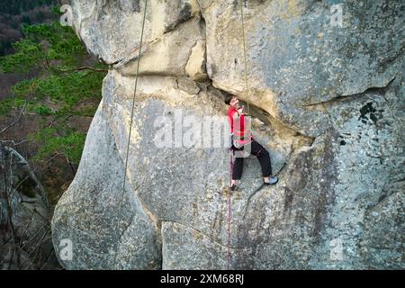 Vue aérienne d'un grimpeur mâle montant une falaise de calcaire accidentée avec harnais et corde pour plus de sécurité. Sportif grimpant sur un grand rocher vertical à Dobvush Rocks dans les montagnes des Carpates, Ukraine. Banque D'Images