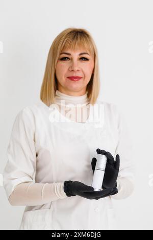 esthéticienne, femme thérapeute beauté portant une blouse de laboratoire blanche tient un produit dans des gants noirs. Elle regarde directement la caméra, souriant légèrement. Banque D'Images