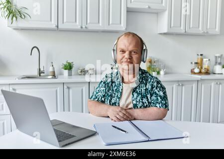 Un homme avec inclusivité portant des écouteurs est assis à une table de cuisine travaillant sur un ordinateur portable et un ordinateur portable. Banque D'Images