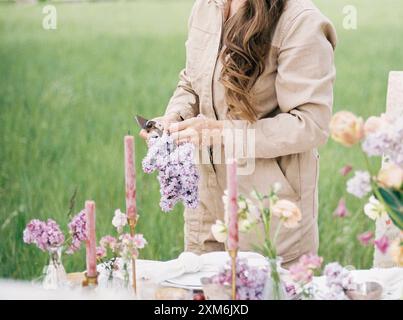 Femme arrangeant des fleurs de lilas sur une table extérieure avec des bougies roses Banque D'Images