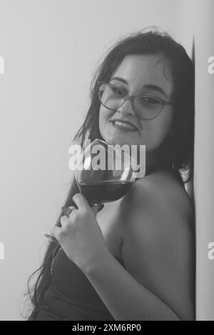 Portrait en noir et blanc d'une femme appuyée contre un mur tenant une tasse de verre avec du vin à l'intérieur. Photo studio. Banque D'Images