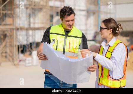 Deux ouvriers de la construction en gilets de sécurité examinant un plan sur un chantier de construction extérieur, collaborant et planifiant des travaux. Banque D'Images