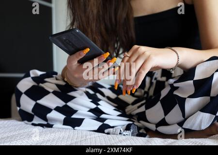 Smartphone dans les mains d'une fille avec des ongles en acrylique orange et de longs cheveux assis sur un canapé. Mode féminine et beauté, communication en ligne Banque D'Images