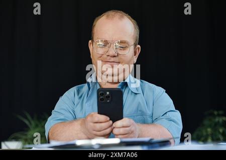Un homme avec inclusivité portant des lunettes utilise son smartphone dans une pièce faiblement éclairée. Banque D'Images