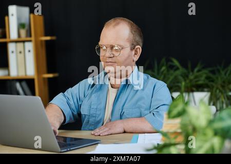 Un homme avec inclusivité dans les lunettes travaille diligemment sur un ordinateur portable à son bureau dans un cadre de bureau. Banque D'Images