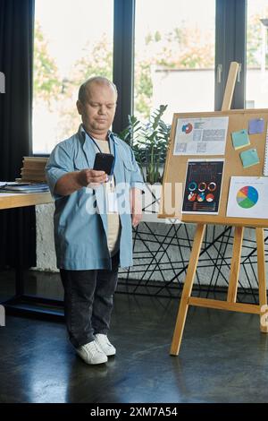 Un homme avec inclusivité vérifie son téléphone tout en se tenant près d'un tableau d'affichage de bureau. Banque D'Images