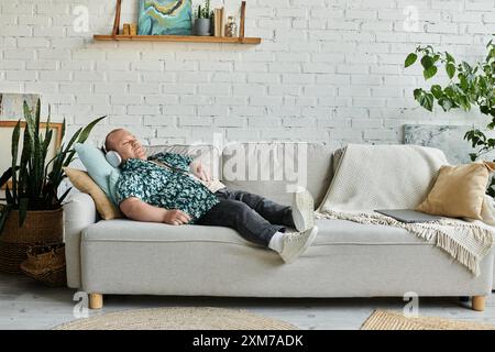 Un homme avec inclusivité repose paisiblement sur un canapé gris, portant des écouteurs et une chemise à motifs. Banque D'Images