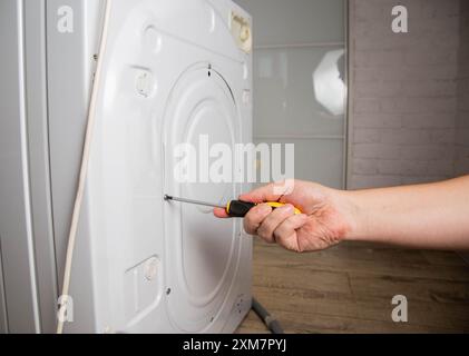 La main d'un maître avec un tournevis dévisse le couvercle arrière de la machine à laver pour le réparer. Banque D'Images