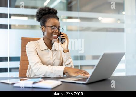 Femme d'affaires souriante dans des lunettes parlant au téléphone tout en travaillant sur ordinateur portable dans un bureau moderne. L'image illustre le professionnalisme, la productivité et l'environnement de travail moderne. Banque D'Images