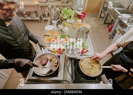 Plan à angle élevé de deux cuisiniers professionnels préparant des pâtes italiennes et un steak angus noir dans une cuisine moderne Banque D'Images