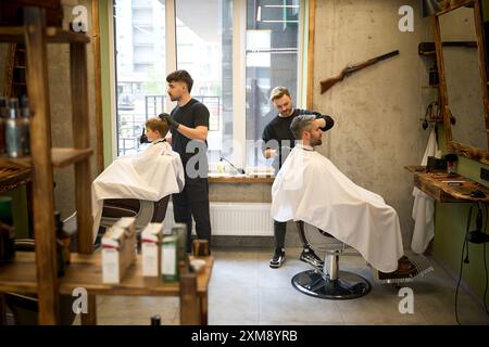 Barbiers qualifiés faisant des coupes de cheveux au père et au fils qui sont assis l'un en face de l'autre Banque D'Images