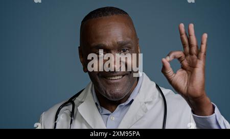 Portrait heureux homme afro-américain souriant médecin thérapeute chirurgien professionnel médical travailleur dans le studio fond bleu montrer OK signe montrant Banque D'Images