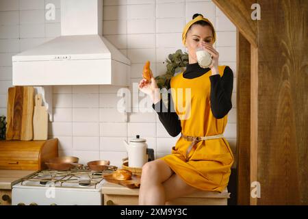 Une femme en robe jaune boit un verre de lait tout en mangeant un pain aux pommes. Elle est assise sur le comptoir de cuisine avec les jambes croisées Banque D'Images