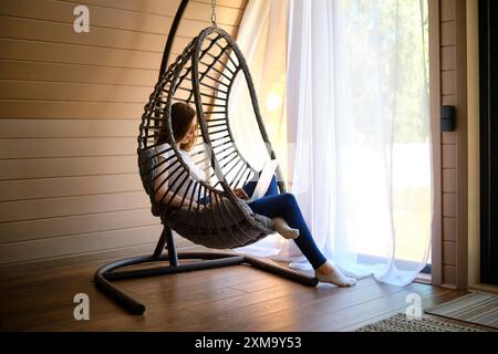 Par une chaude journée ensoleillée, une jeune femme est assise dans une chaise pendulaire avec un ordinateur portable dans une pièce climatisée de la cabane en rondins Banque D'Images