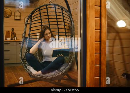 Une jeune femme se détend le soir dans une maison en bois assise dans une chaise suspendue avec un livre Banque D'Images