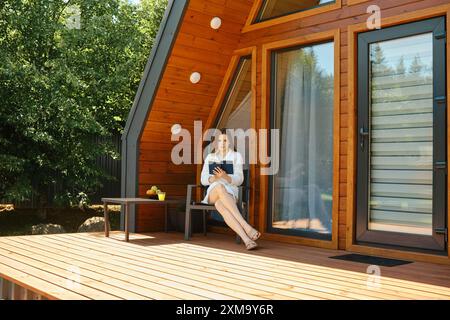 Pendant les soins cosmétiques quotidiens de la peau du visage, une femme en peignoir lit un livre par un matin chaud et ensoleillé sur la terrasse d'une maison en bois Banque D'Images