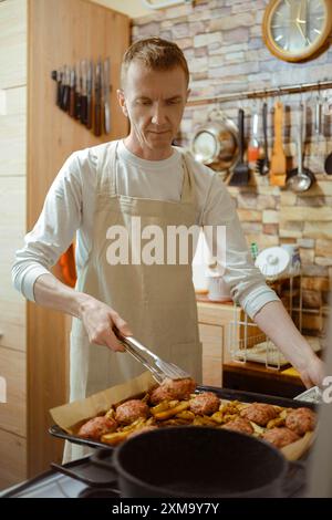 L'homme d'âge moyen dans un tablier transfère les escalopes de viande d'une plaque de cuisson à une casserole à l'aide d'une pince de cuisine Banque D'Images