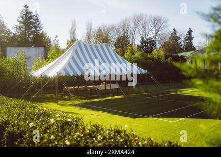 Une tente rayée bleue et blanche dans le jardin, entourée d'arbres, avec des tables et un ciel ensoleillé, Hamilton, Nouvelle-Zélande Banque D'Images