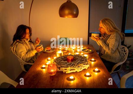 Image symbolique économie d'énergie, hiver froid, crise énergétique, plat froid, mère et fille vêtues chaudement au dîner, seulement à la lueur des chandelles, chauffage sur Banque D'Images