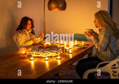 Image symbolique économie d'énergie, hiver froid, crise énergétique, plat froid, mère et fille vêtues chaudement au dîner, seulement à la lueur des chandelles, chauffage sur Banque D'Images