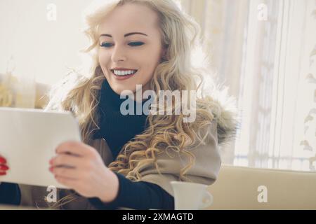 Jeune femme aux longs cheveux blonds bouclés sourit tout en utilisant une tablette dans un café confortable, mélangeant technologie et détente dans un cadre élégant Banque D'Images